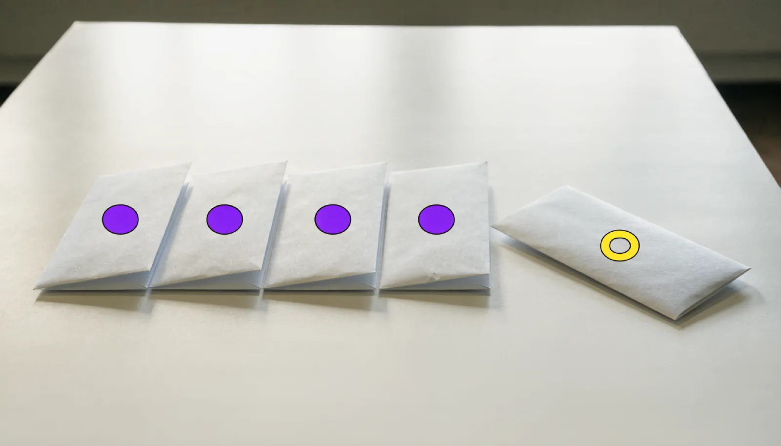 A photograph of five plain envelopes lying in a row on a table, each marked with a small purple dot, and a sixth envelope set apart on its own marked with a yellow ring.